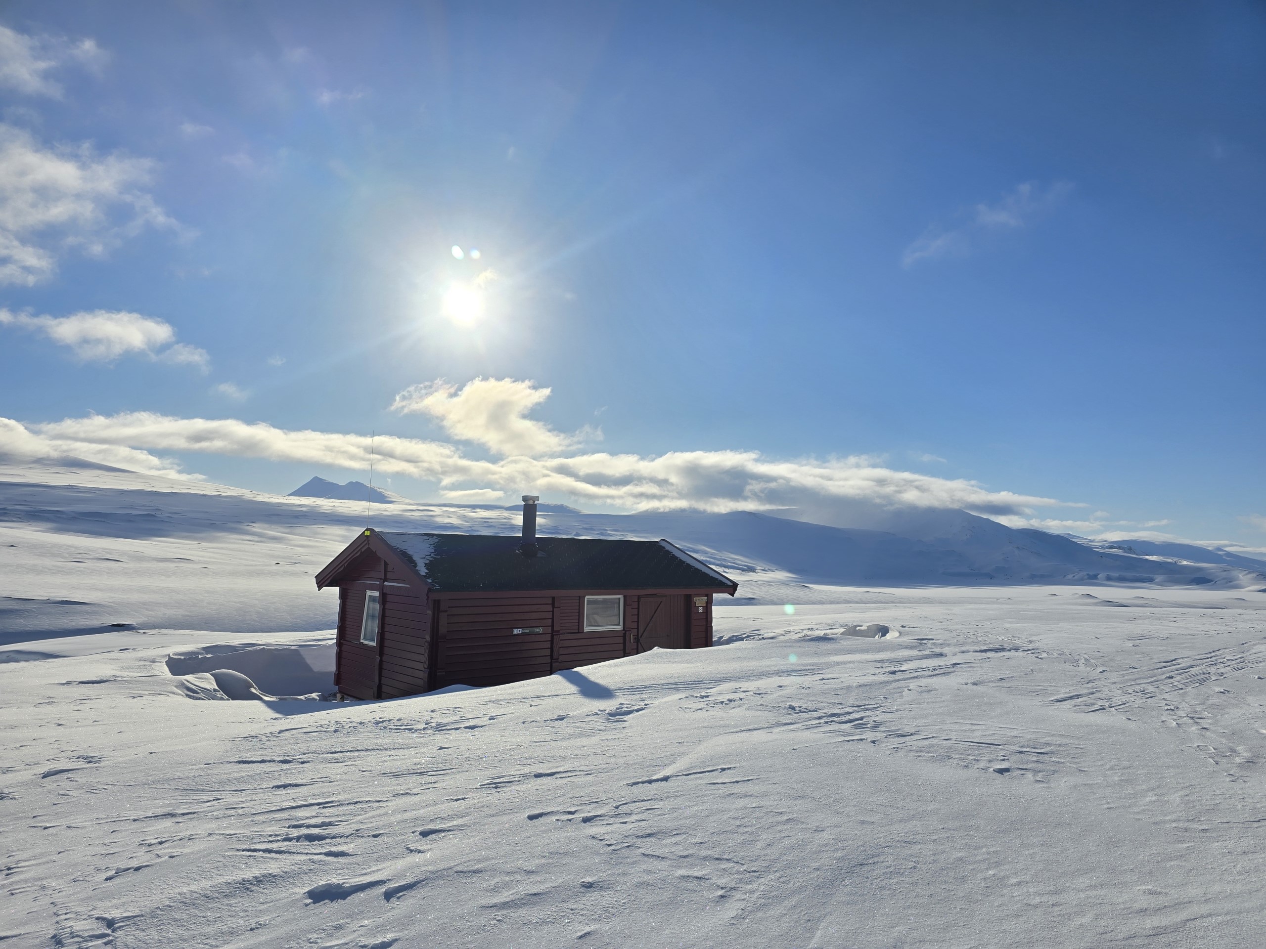Ei rødbrun hytte med svart tak. Hytta ligger på høyfjellet. Mye snø på bildet.